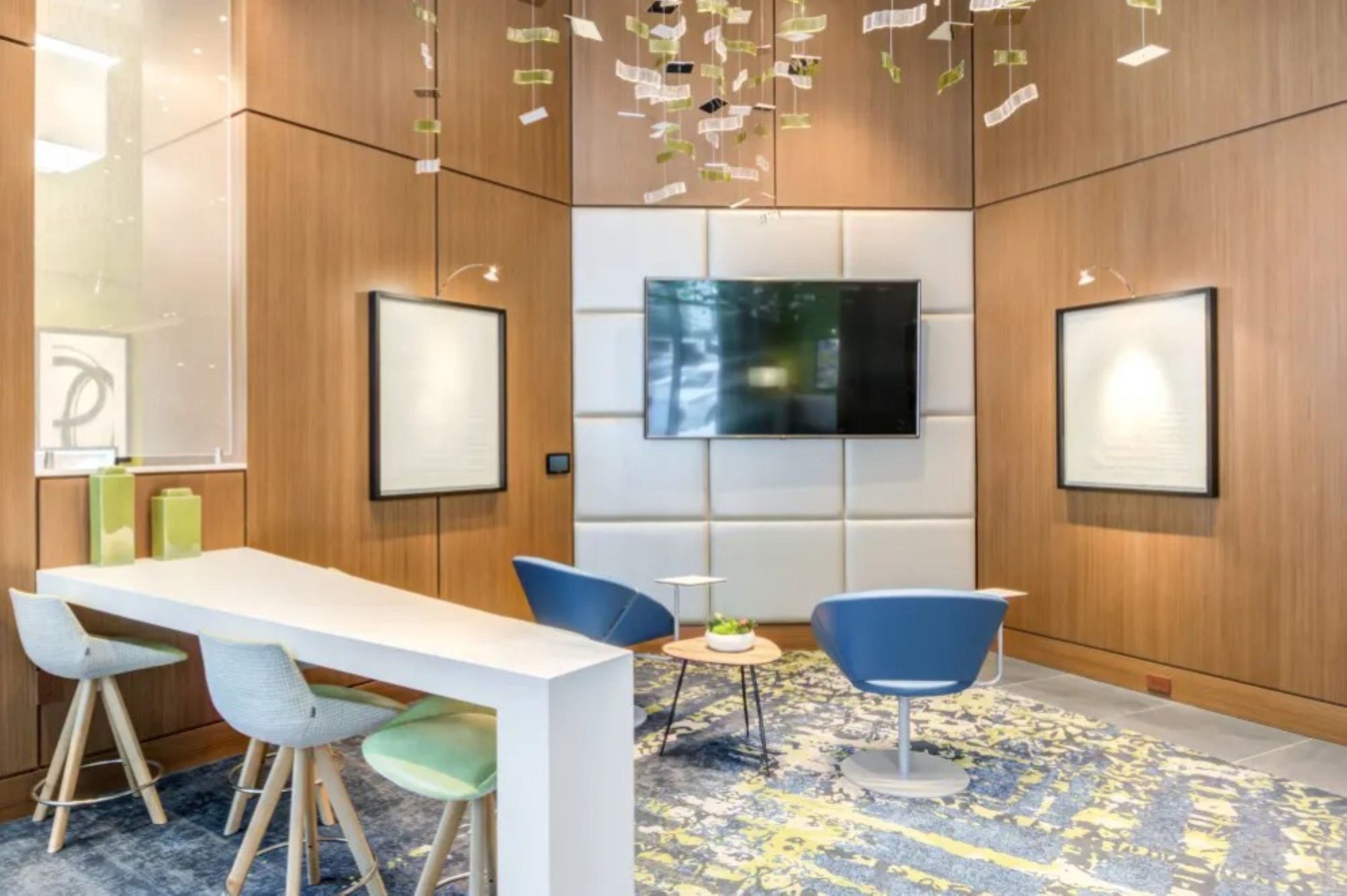 Una sala de conferencias moderna con una mesa blanca, sillas azules, un televisor montado y paneles de madera.