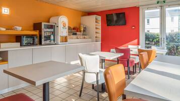 Un café moderne avec des chaises blanches et orange, une machine à café et un téléviseur fixé au mur.
