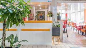 A reception area with a modern design, featuring a peacock feather artwork, potted plants, and a seating area with orange chairs and tables. - PREMIERE CLASSE ROUEN NORD - Barentin (Barentin)