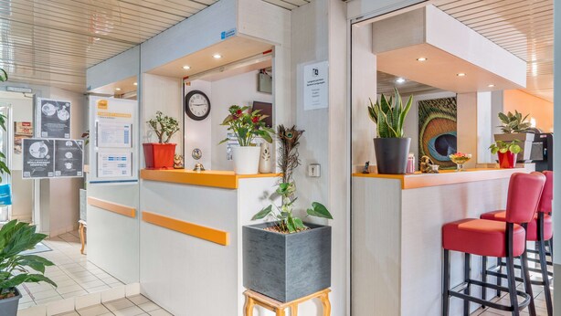 A reception area with a modern design, featuring a reception desk, a clock, and various potted plants. - PREMIERE CLASSE ROUEN NORD - Barentin (Barentin)