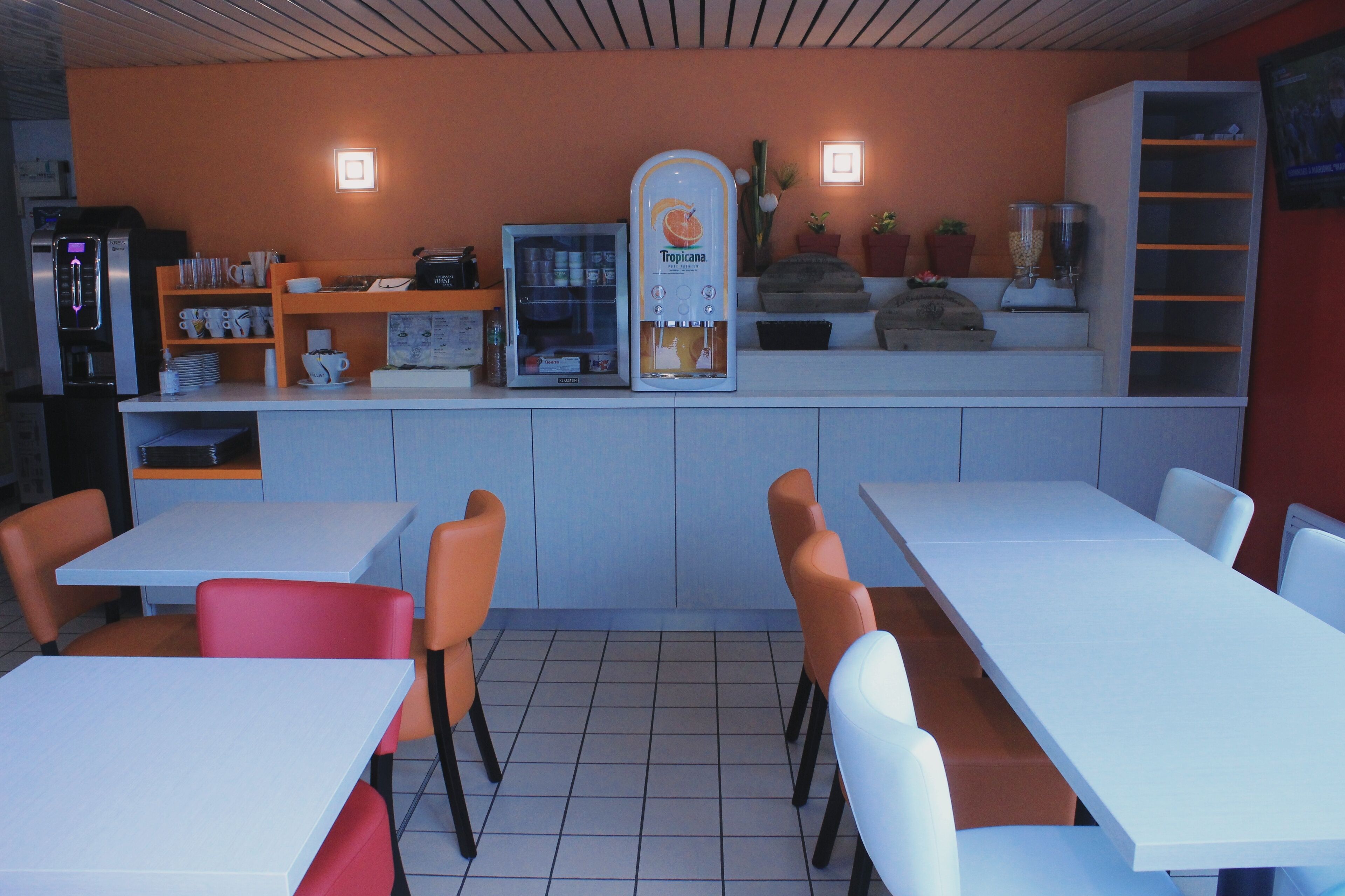 A restaurant with white tables and orange chairs, a coffee machine, and a refrigerator.