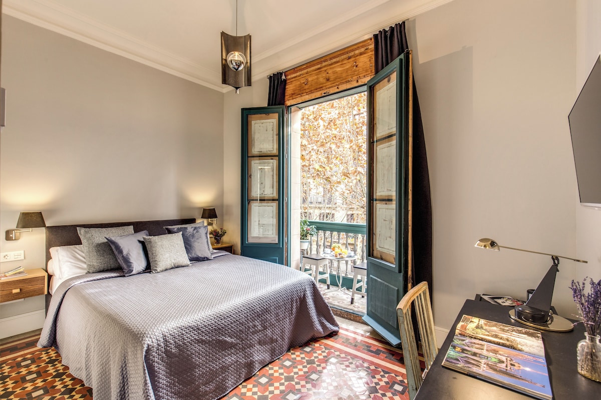 A bedroom with a large bed, a wooden door leading to a balcony, a desk with a lamp, and a view of trees outside.