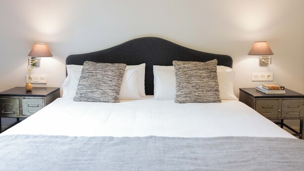 A neatly made bed with white linens and two patterned pillows, flanked by two bedside tables with lamps and a small book on one. - We Boutique Hotel (Barcelona)
