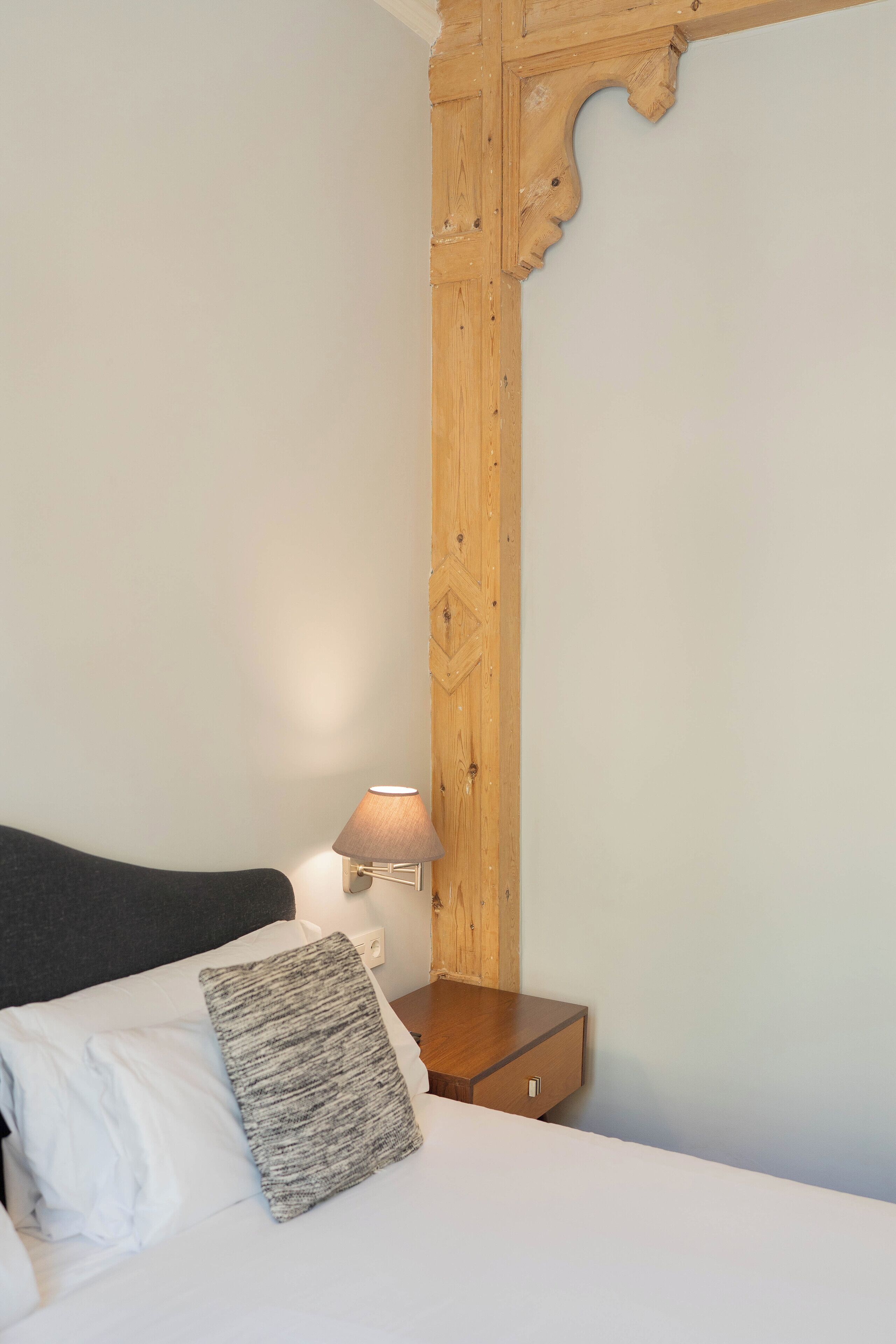 A neatly made bed with white linens and a grey pillow, a wooden nightstand with a lamp, and a wooden wall panel.