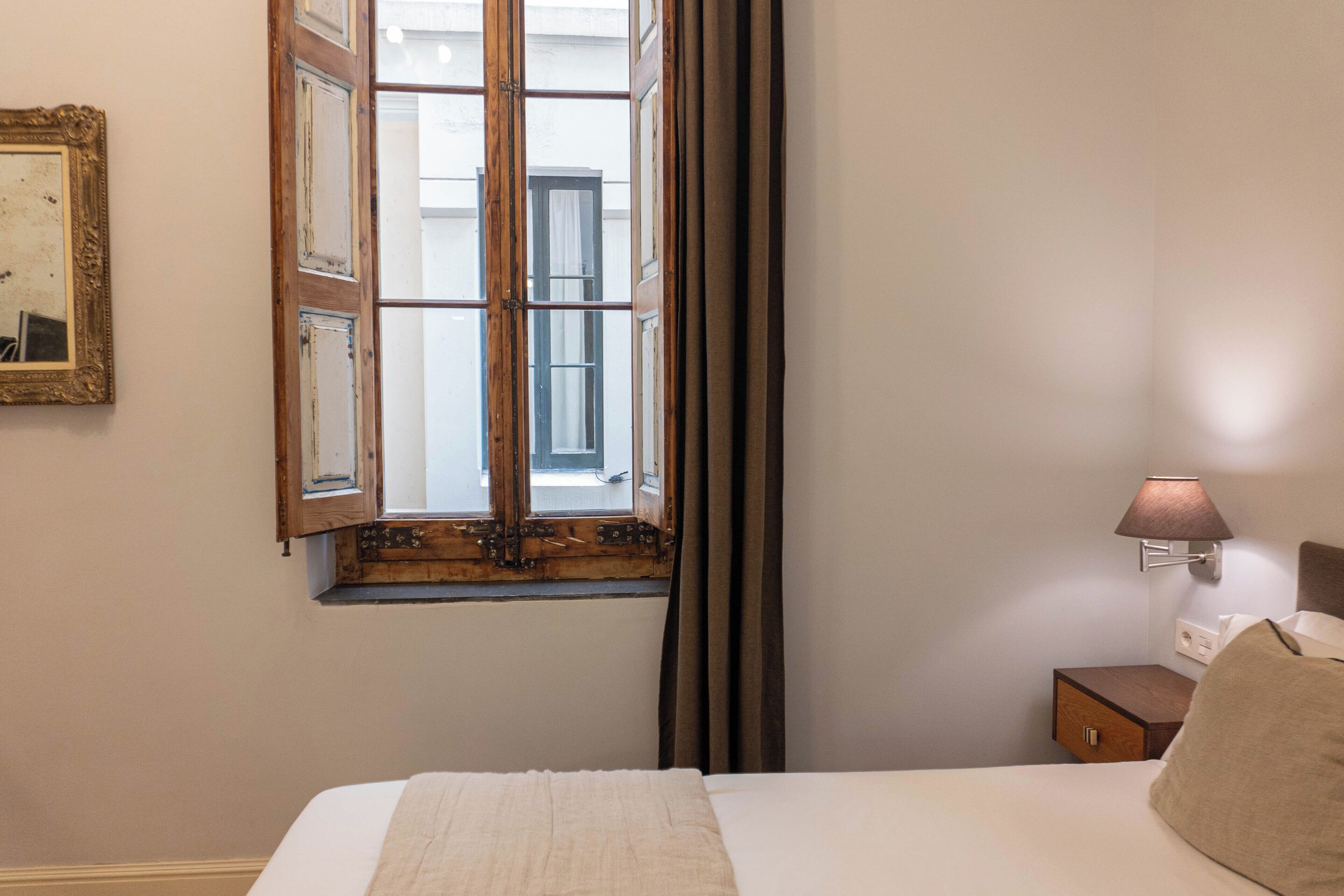 Un dormitorio con cama, mesita de noche, lámpara, ventana con cortinas y un cuadro en la pared.