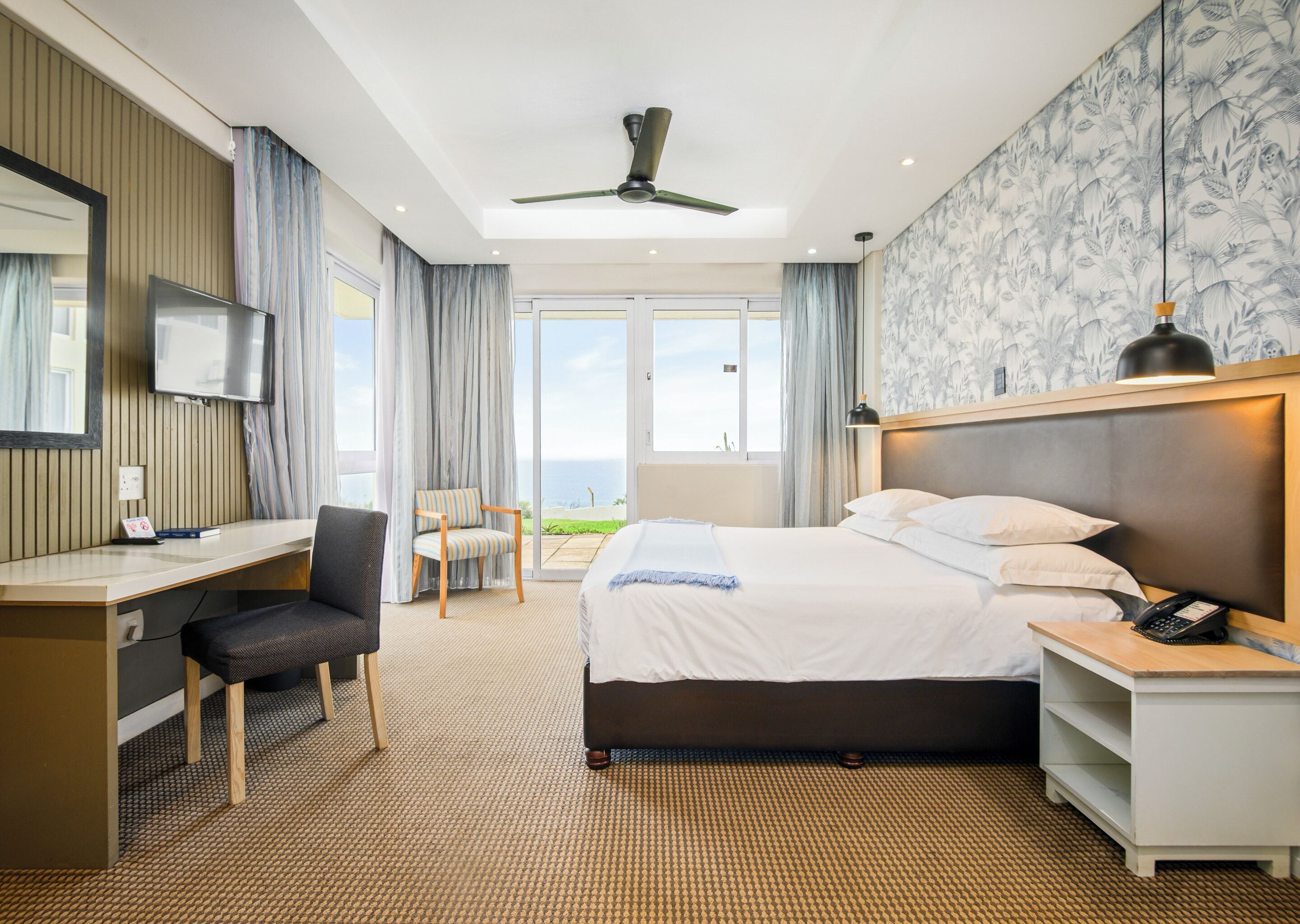A hotel room with a bed, desk, chair, and a view of the sea.