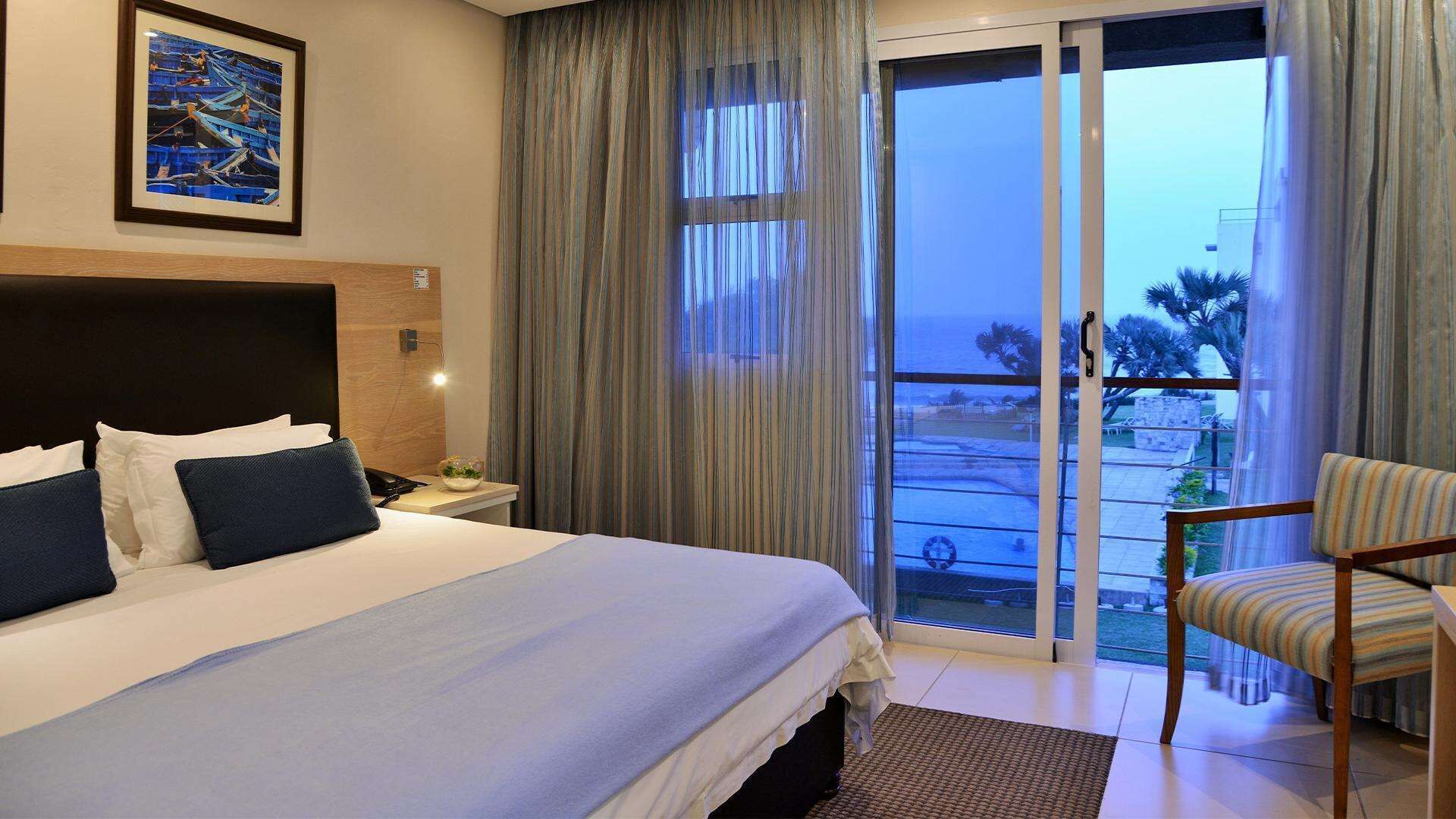 A hotel room with a large bed, a nightstand, a chair, and a view of a pool and palm trees through a sliding glass door.