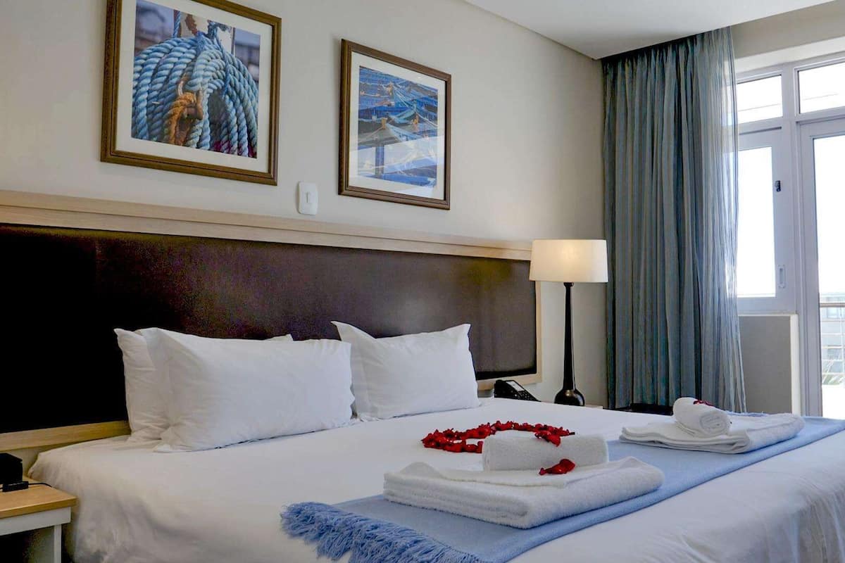 Habitación de hotel con una cama grande, dos cuadros en la pared, una lámpara de noche y vistas al exterior a través de una ventana.