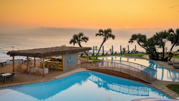 A pool area with a thatched-roof structure, palm trees, and a view of the ocean at sunset. - Blue Marlin All-Inclusive Seascape by Dream Resorts (Scottburgh)