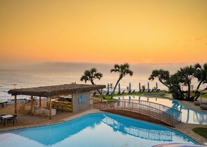 A pool area with a thatched-roof structure, palm trees, and a view of the ocean at sunset. - Blue Marlin All-Inclusive Seascape by Dream Resorts (Scottburgh)