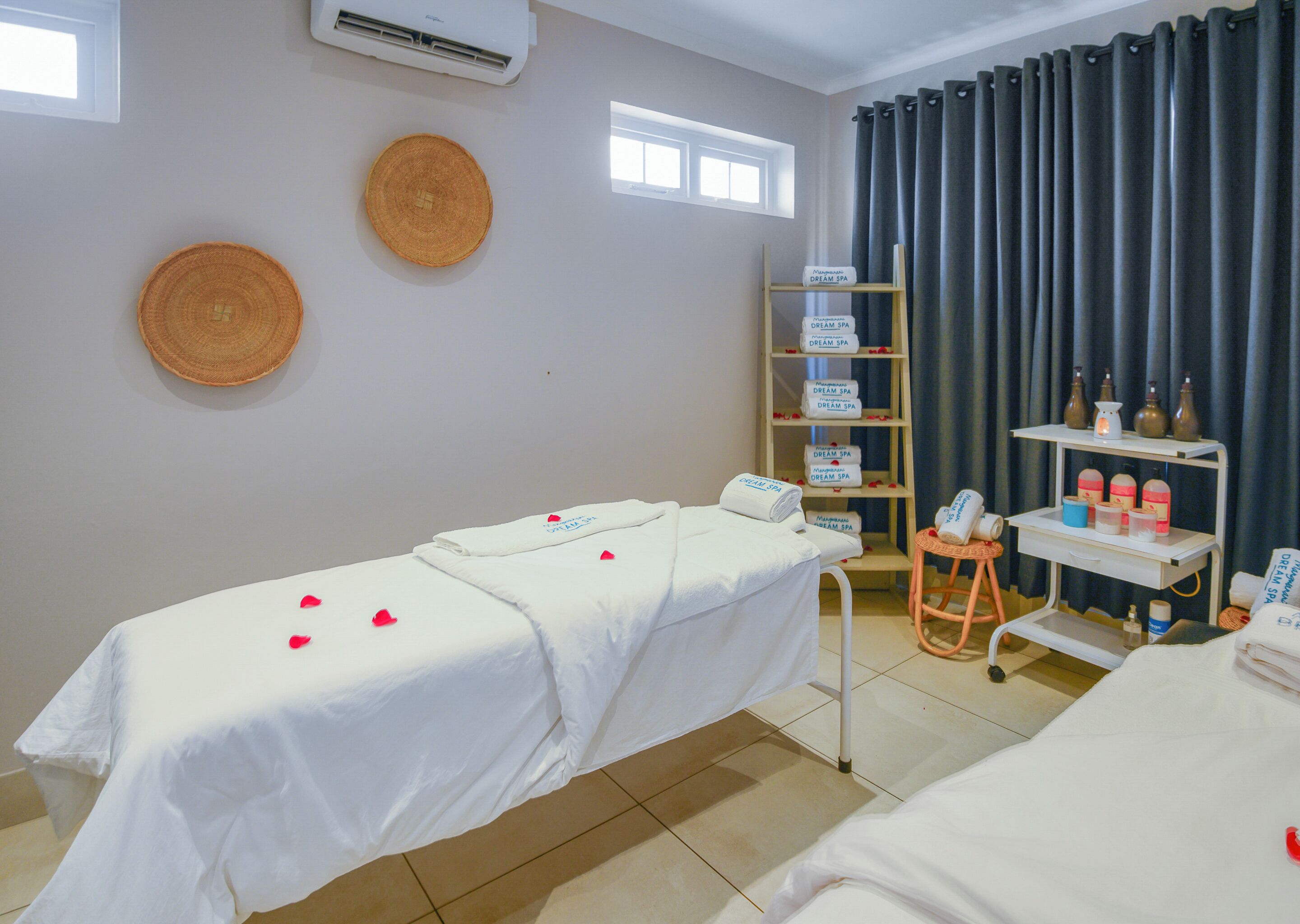 A spa room with a massage table, towels, a shelf with spa products, and a window with curtains.