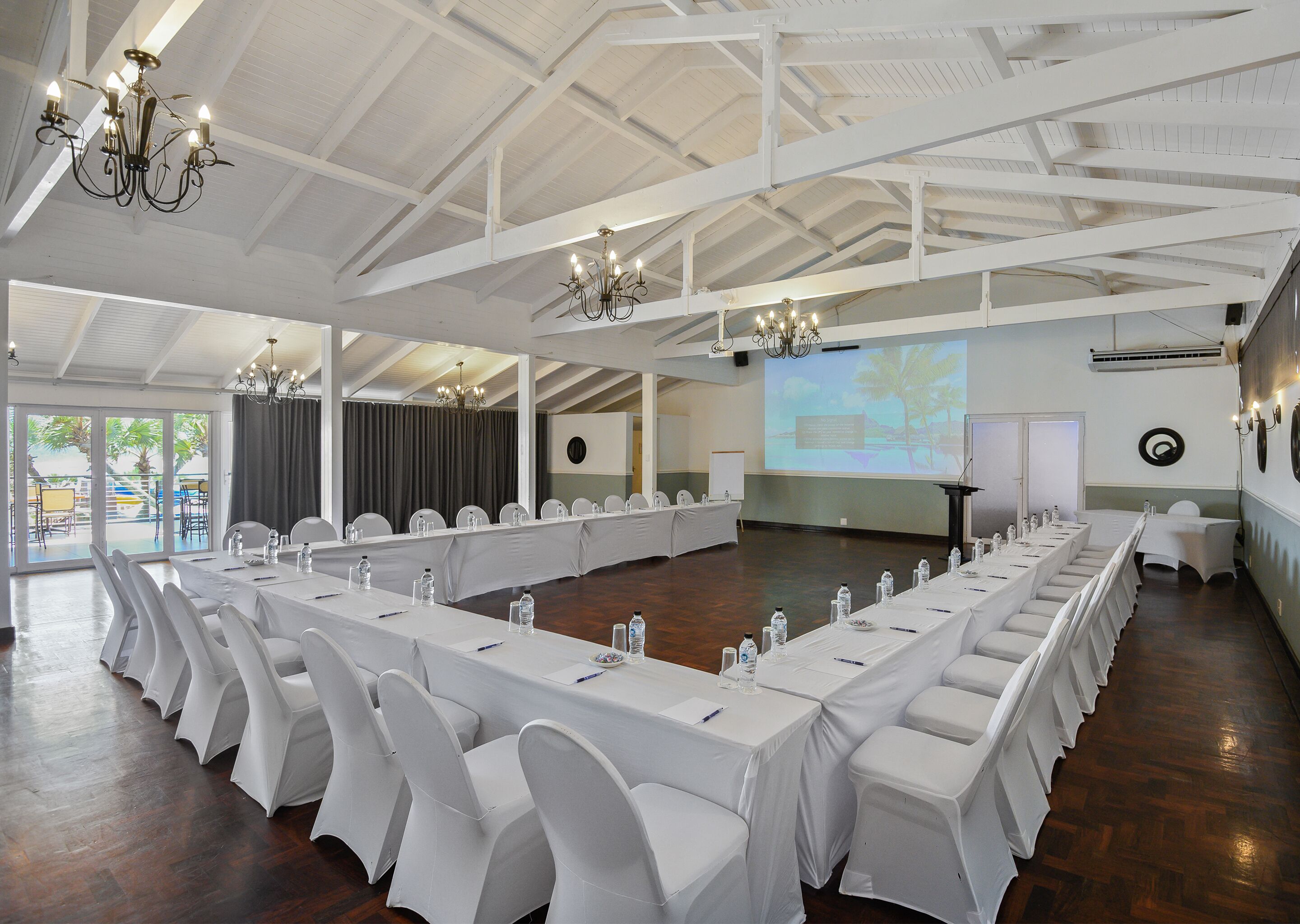 Une salle de conférence avec de longues tables, des chaises blanches, des lustres et un écran de projection.