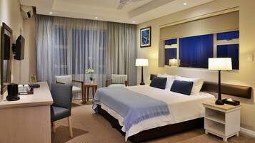 Ein Hotelzimmer mit Bett, Nachttischen, Schreibtisch, Sessel, Lampe, einem Gemälde und einem Fenster mit Vorhängen.