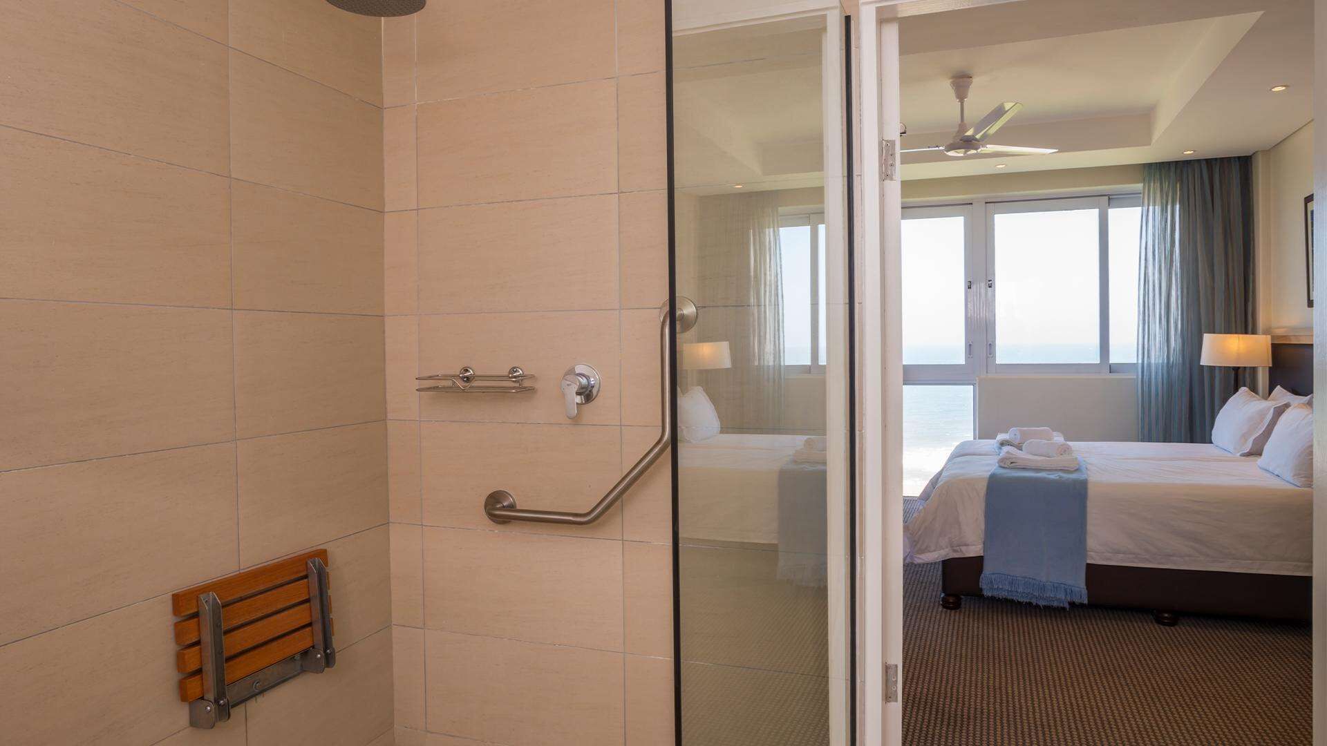 Een hotelkamer met een glazen douchedeur, een houten bankje, een bed met witte lakens en uitzicht op zee.