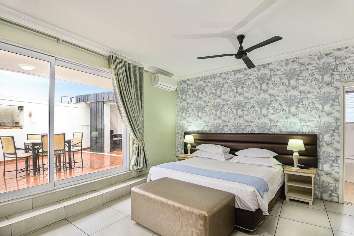 Habitación de hotel con cama, ventilador de techo, mesita de noche, lámpara, ventana con cortinas y vistas al exterior.