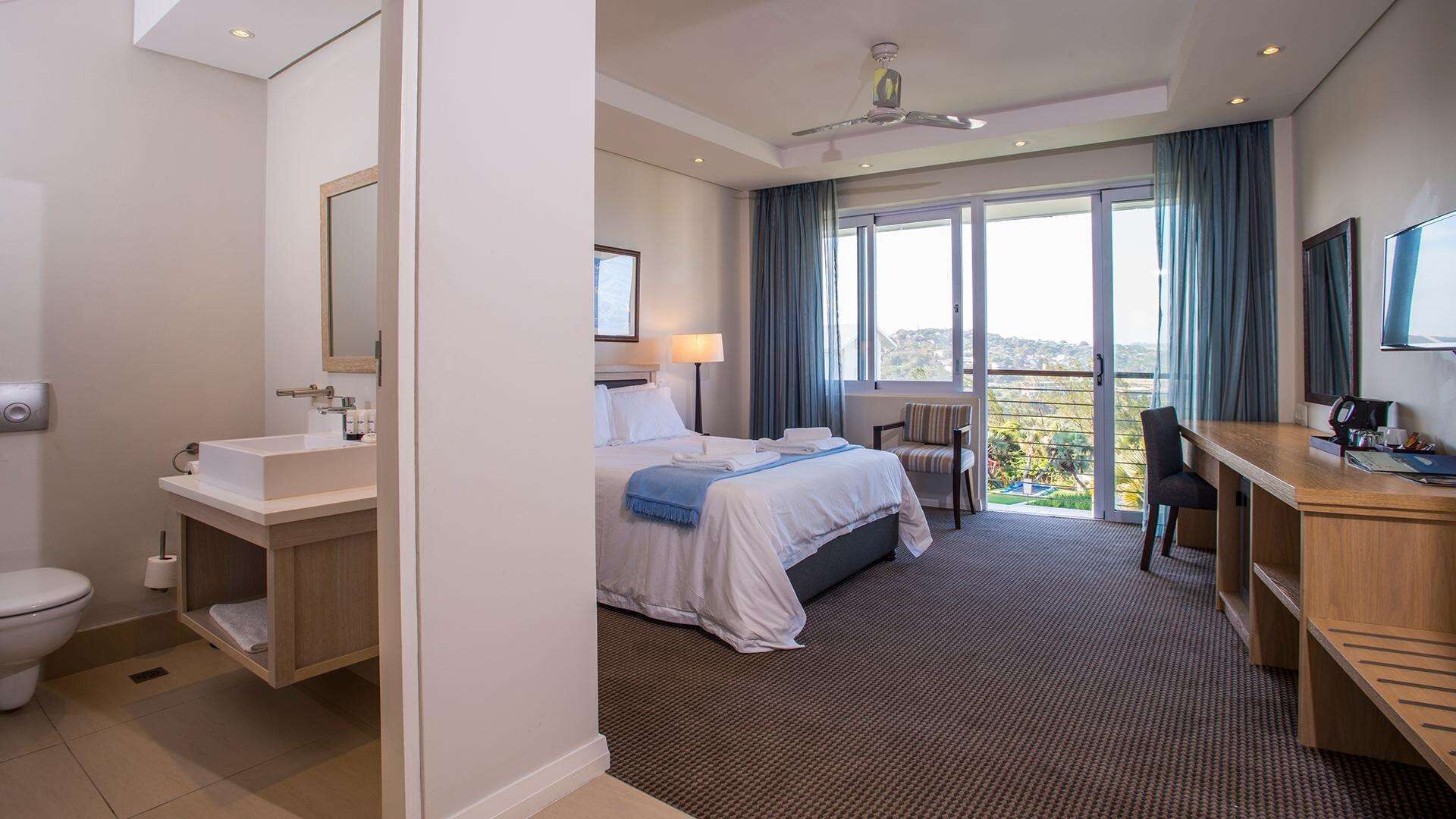 A hotel room with a bed, a desk, a chair, a bathroom, and a balcony with a view.