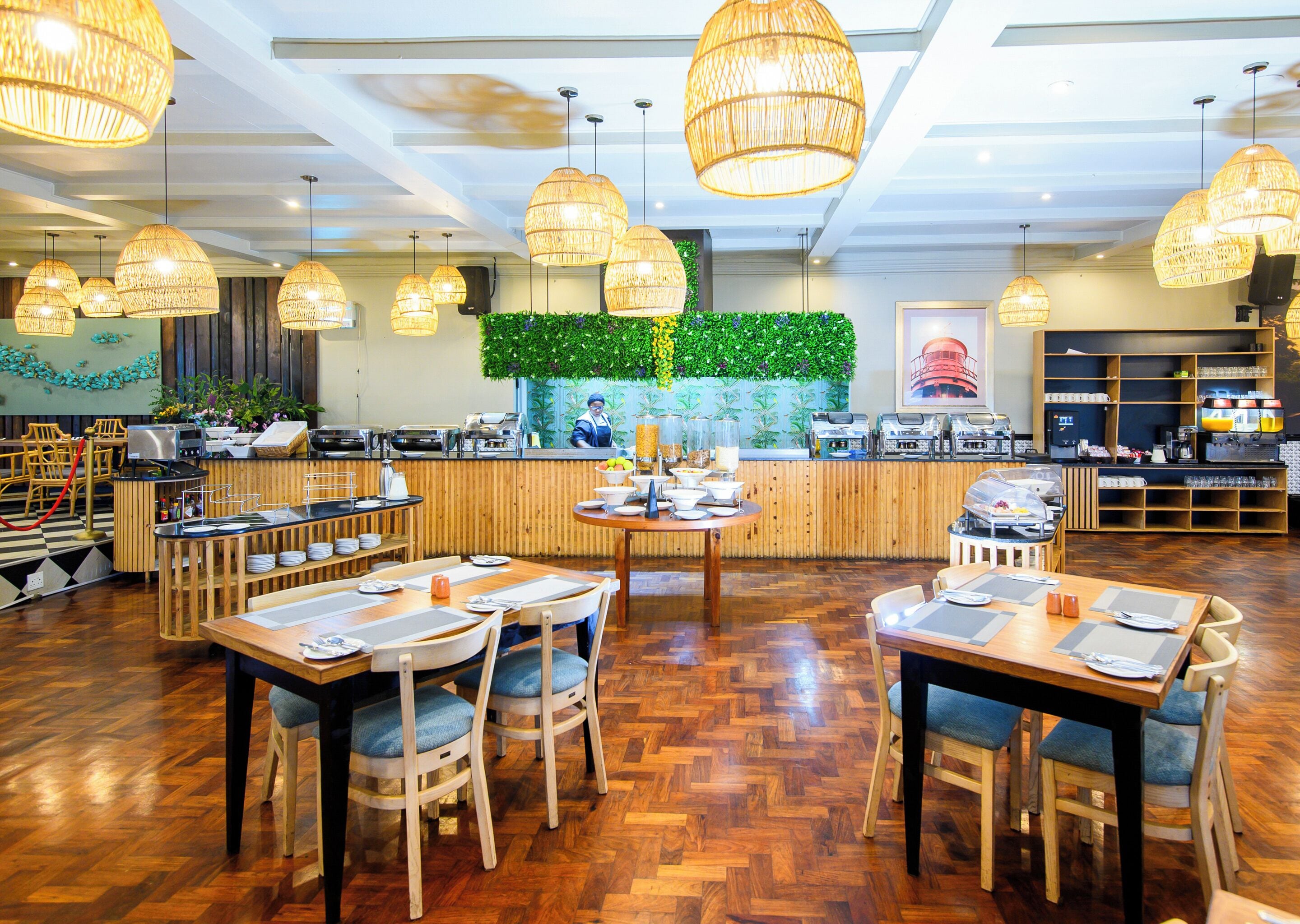 A restaurant with wooden flooring, round tables, and chairs. There are hanging lights and a counter with a chef.