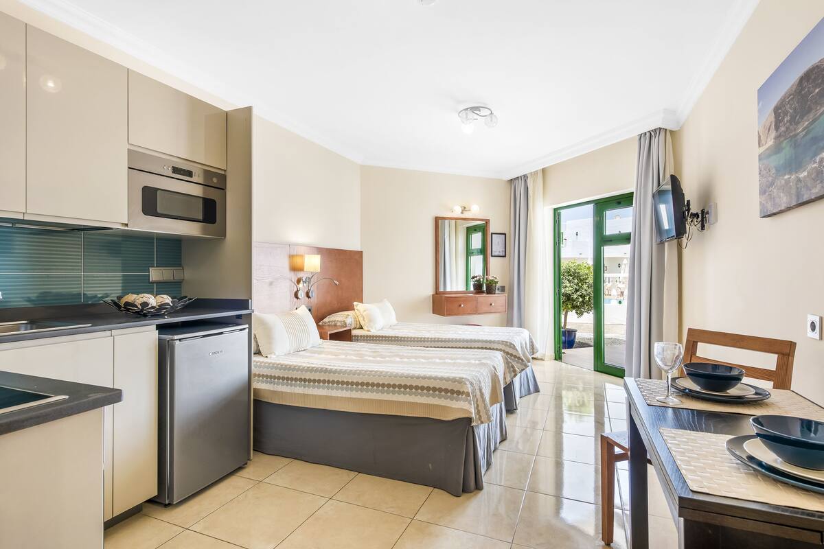 A compact hotel room with a kitchenette, two beds, a dining area, and a view of the outdoors.