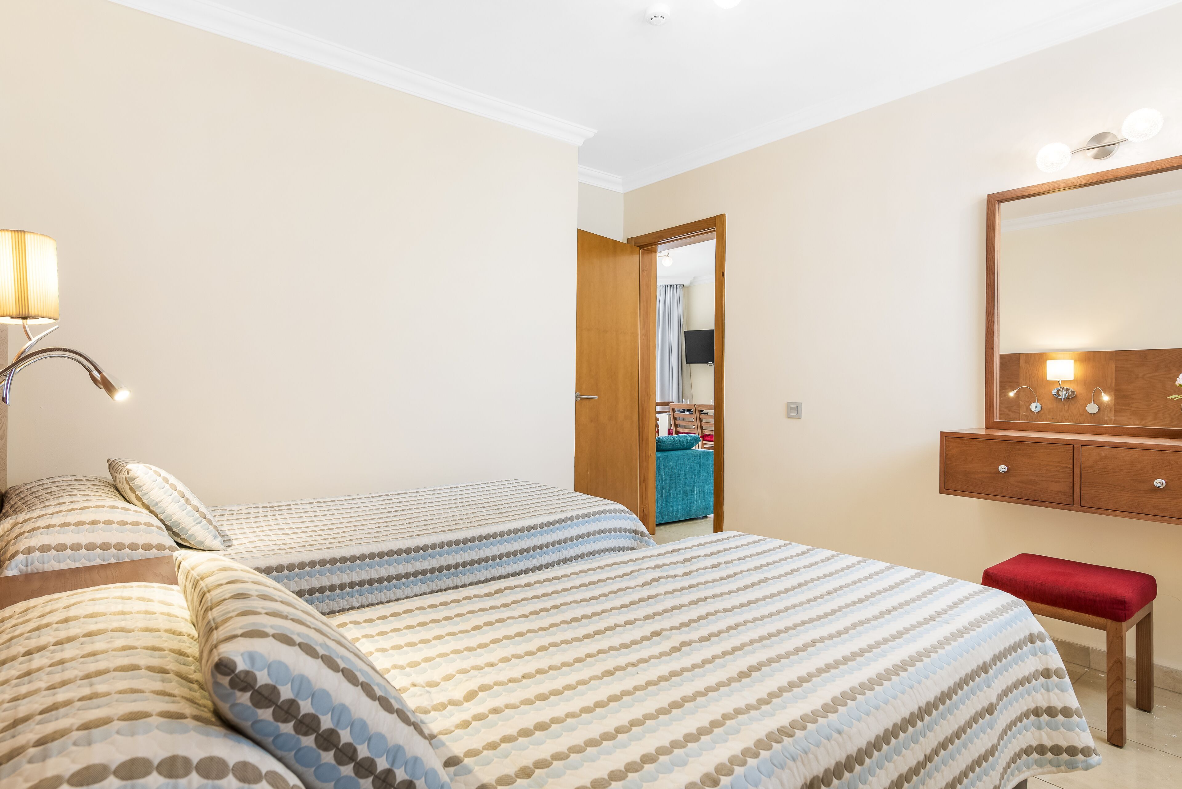 A hotel room with a large bed, a wooden bench, a mirror, and a door leading to a bathroom.