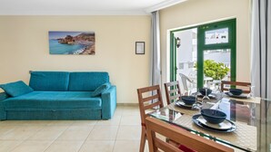 Un salon avec un canapé de couleur turquoise, une table à manger avec des chaises, un tableau représentant un paysage côtier et une vue à travers une porte verte.