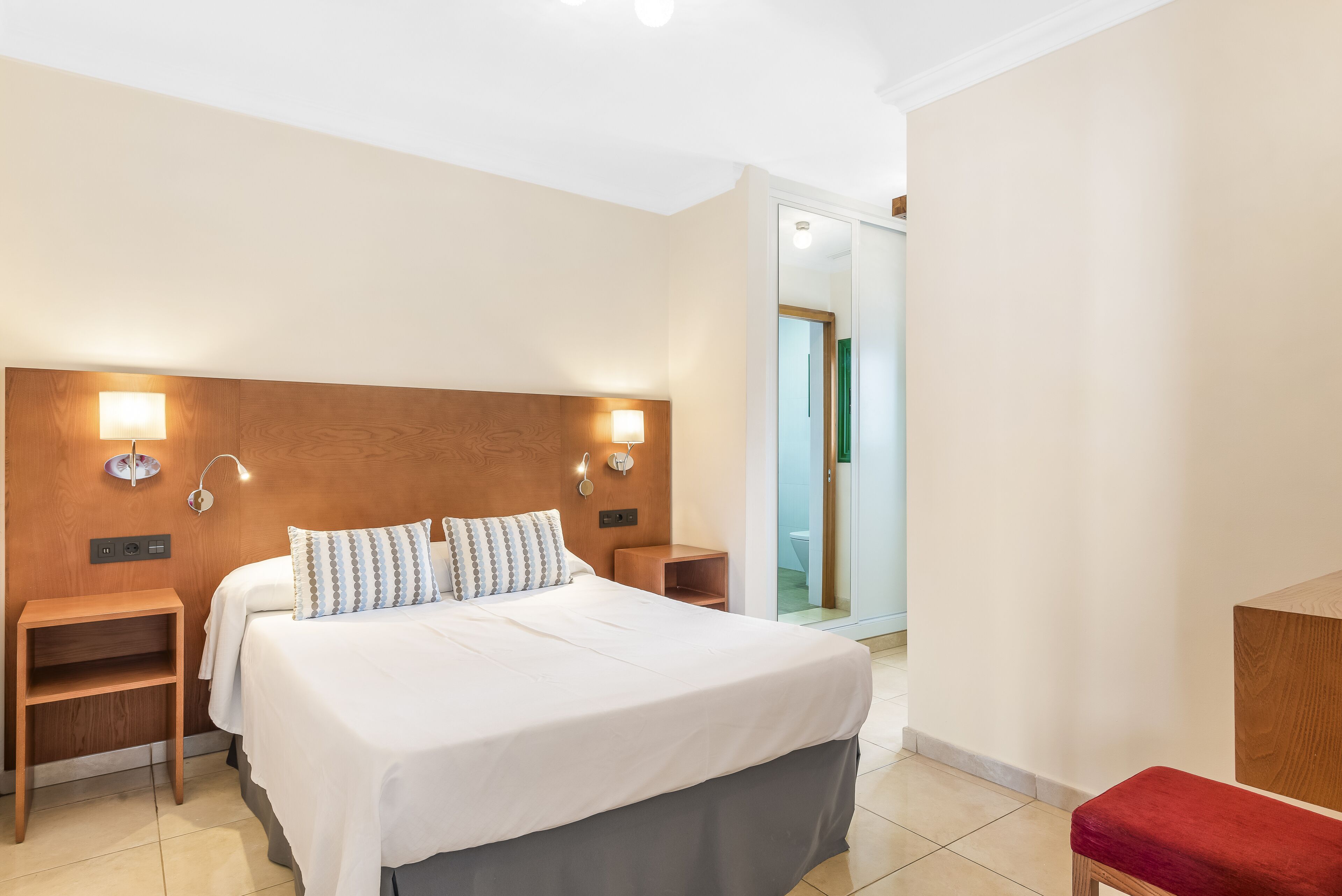 Een hotelkamer met een groot bed, een houten hoofdeinde, bedlampjes, een klein houten nachtkastje en een rode bank.