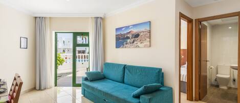 Ein Wohnzimmer mit einem petrolfarbenen Sofa, einem Esstisch mit Stühlen, einem großen Fenster mit Vorhängen und einem durch eine offene Tür einsehbaren Badezimmer.