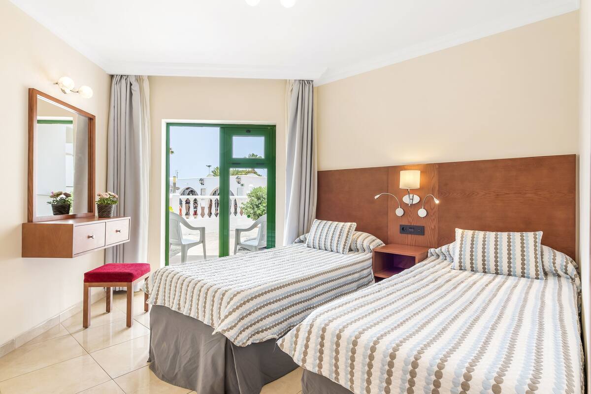 A hotel room with two beds, a desk, a chair, a small stool, and a window with a view of the outside.