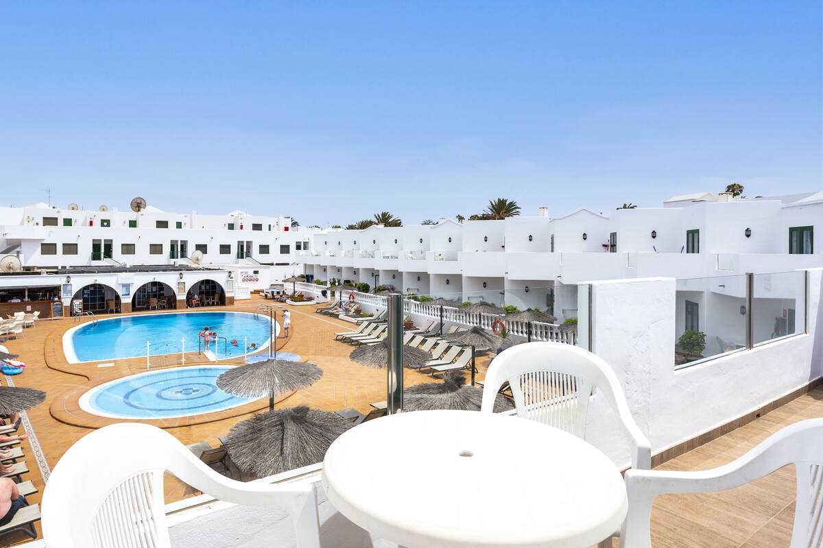 A rooftop pool area with lounge chairs and umbrellas, surrounded by white buildings with balconies.