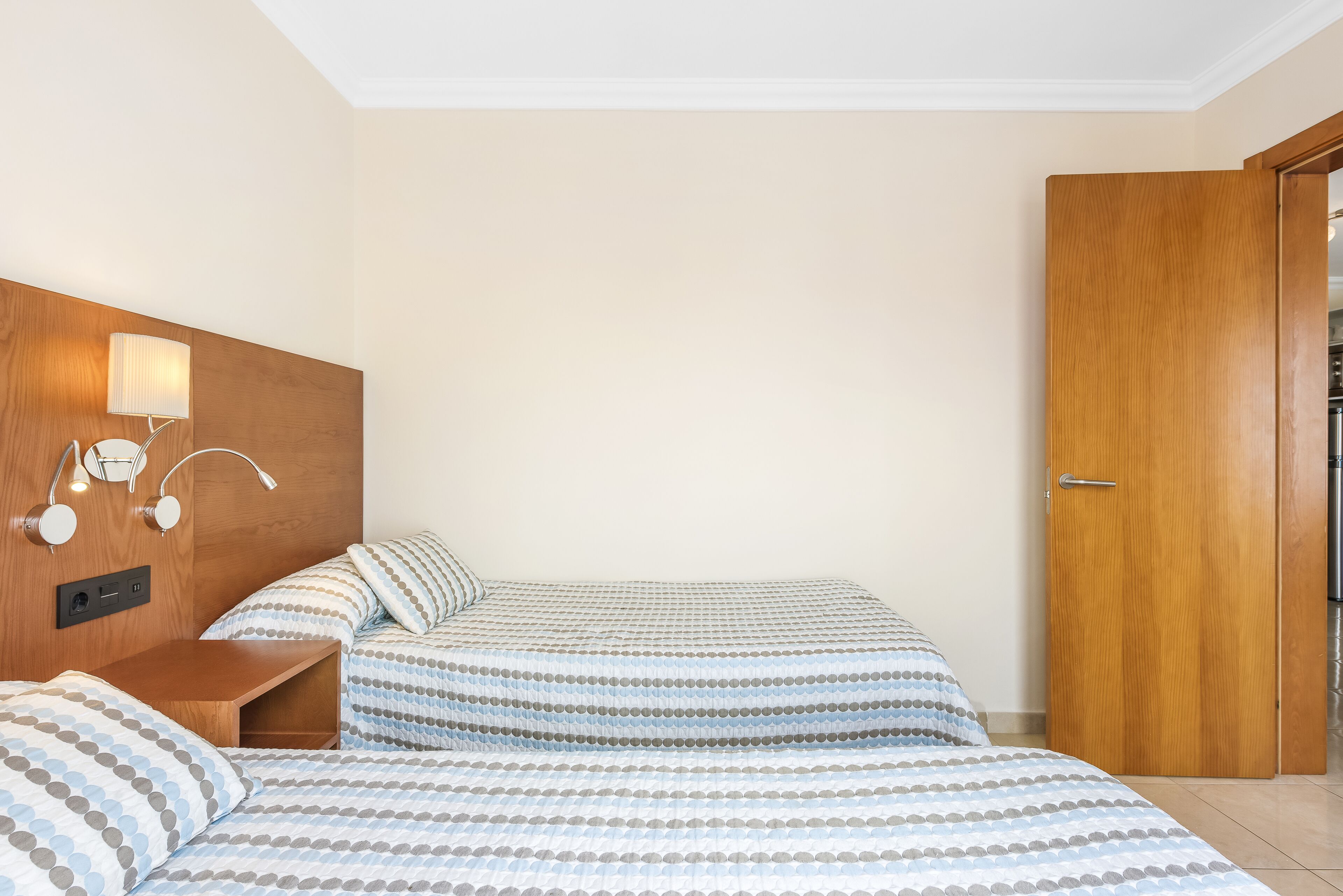Habitación de hotel con dos camas, cabecero de madera, mesita de noche y lámpara de pared.
