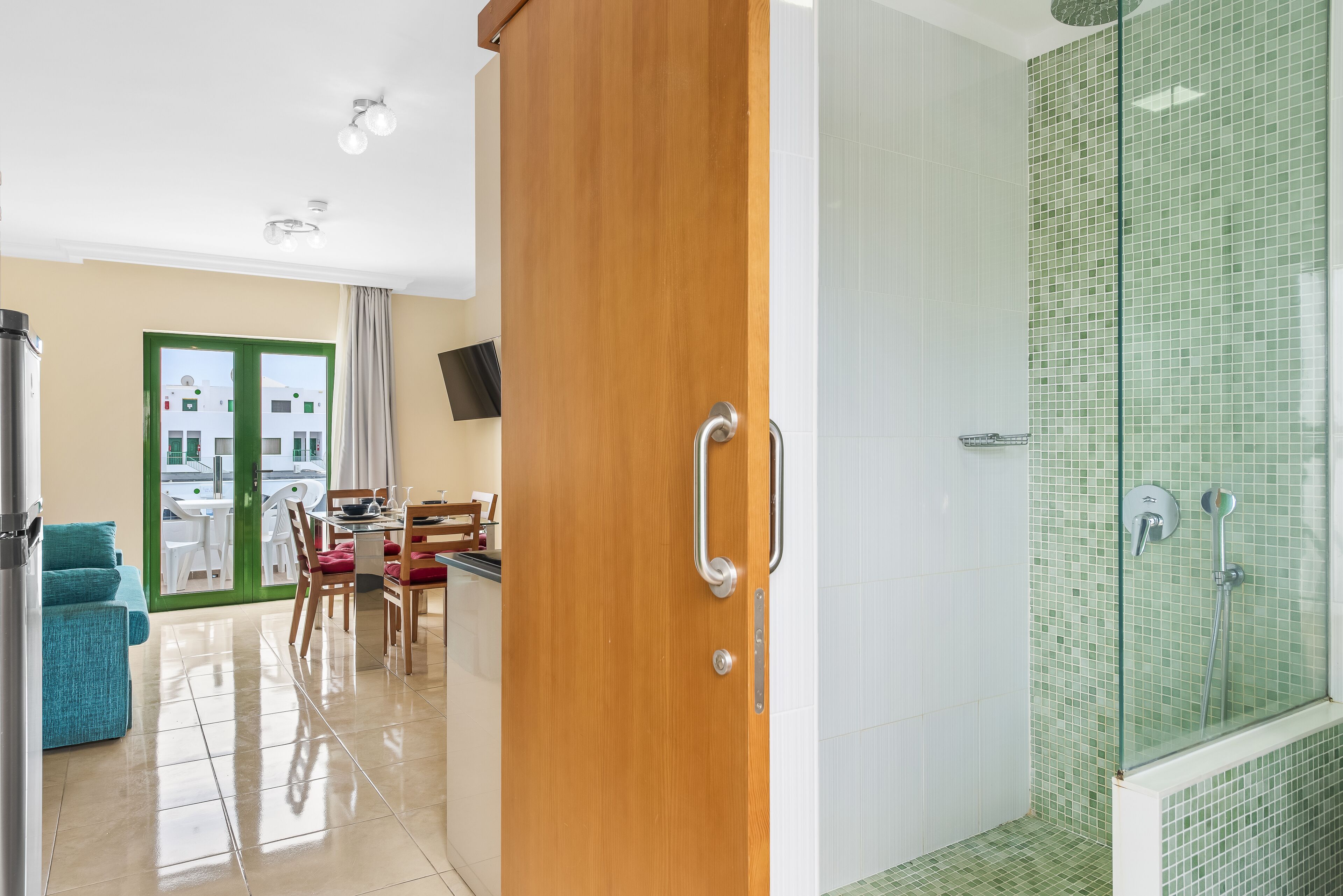 A modern hotel room with a wooden door, a shower with green mosaic tiles, a dining area with a table and chairs, and a view of the outside through a glass door.