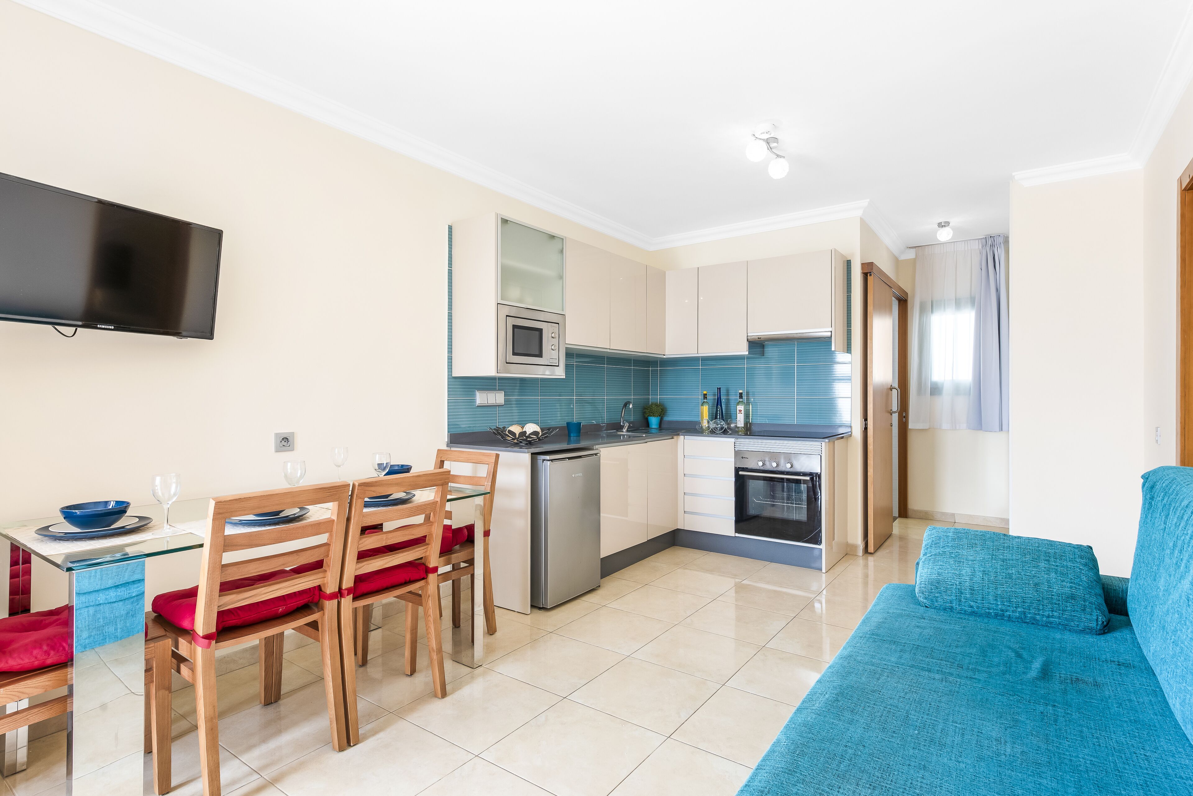 Una cocina moderna con un televisor de pantalla plana, una zona de comedor con sillas de madera y una mesa de vidrio, y un sofá azul en la sala de estar.