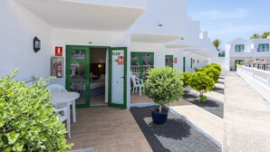 Un bâtiment blanc avec des portes vertes et des chambres numérotées, des places assises à l’extérieur et des plantes en pot.