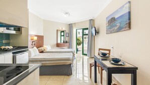 Une chambre d’hôtel moderne équipée d’une kitchenette, d’un lit avec deux oreillers, d’une table à manger avec un bol et un verre, et d’un tableau représentant un paysage côtier.