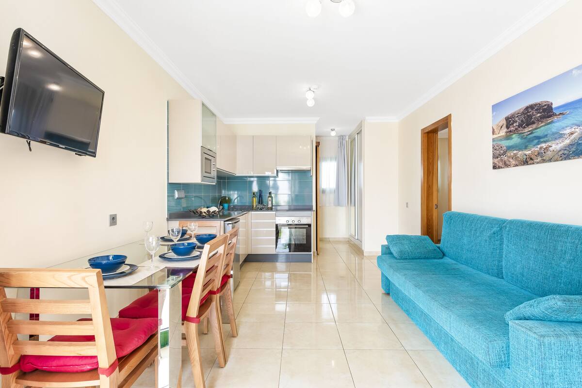 A modern kitchen with a flat-screen TV, a dining area with a glass table, and a blue sofa.
