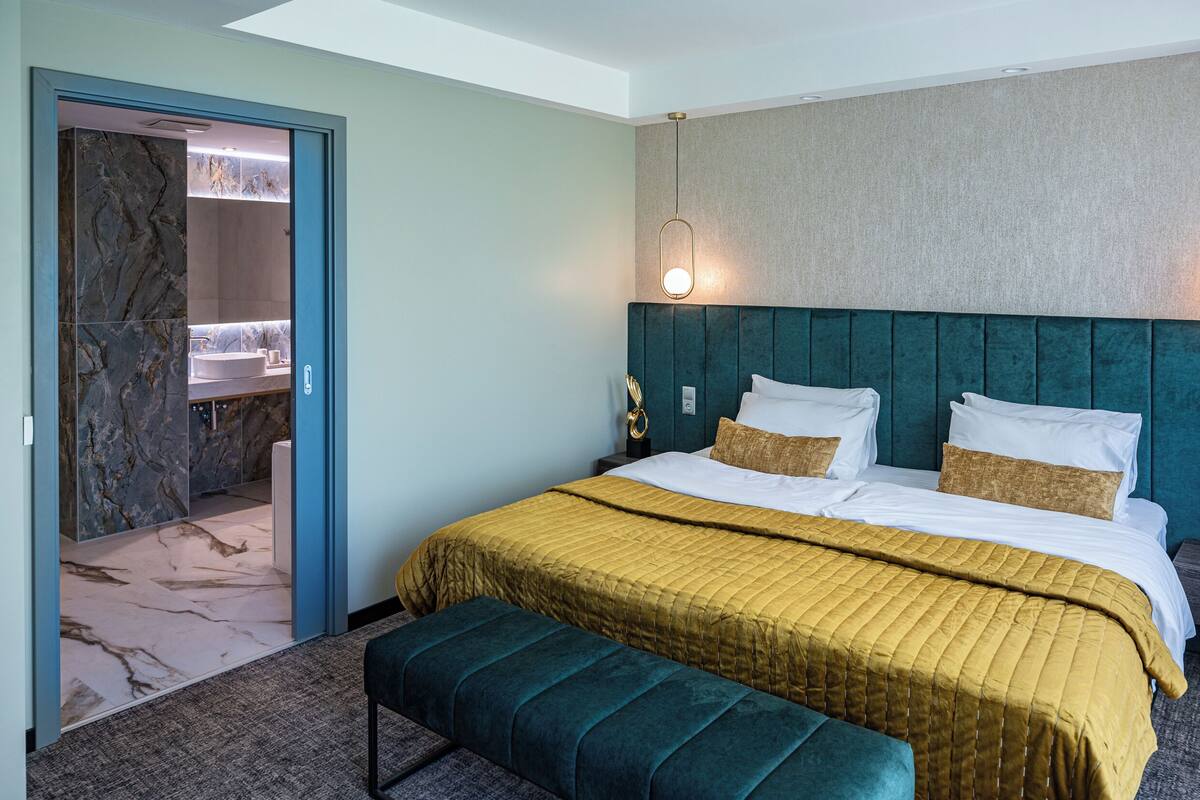A modern hotel room with a large bed, a bench, a bathroom with a marble wall, and a wall-mounted light fixture.