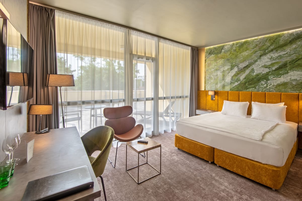 A hotel room with a large bed, a desk with a lamp, a chair, and a view of the outdoors through large windows.