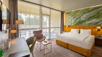 A hotel room with a large bed, a desk with a lamp, a chair, and a view of the outdoors through large windows.