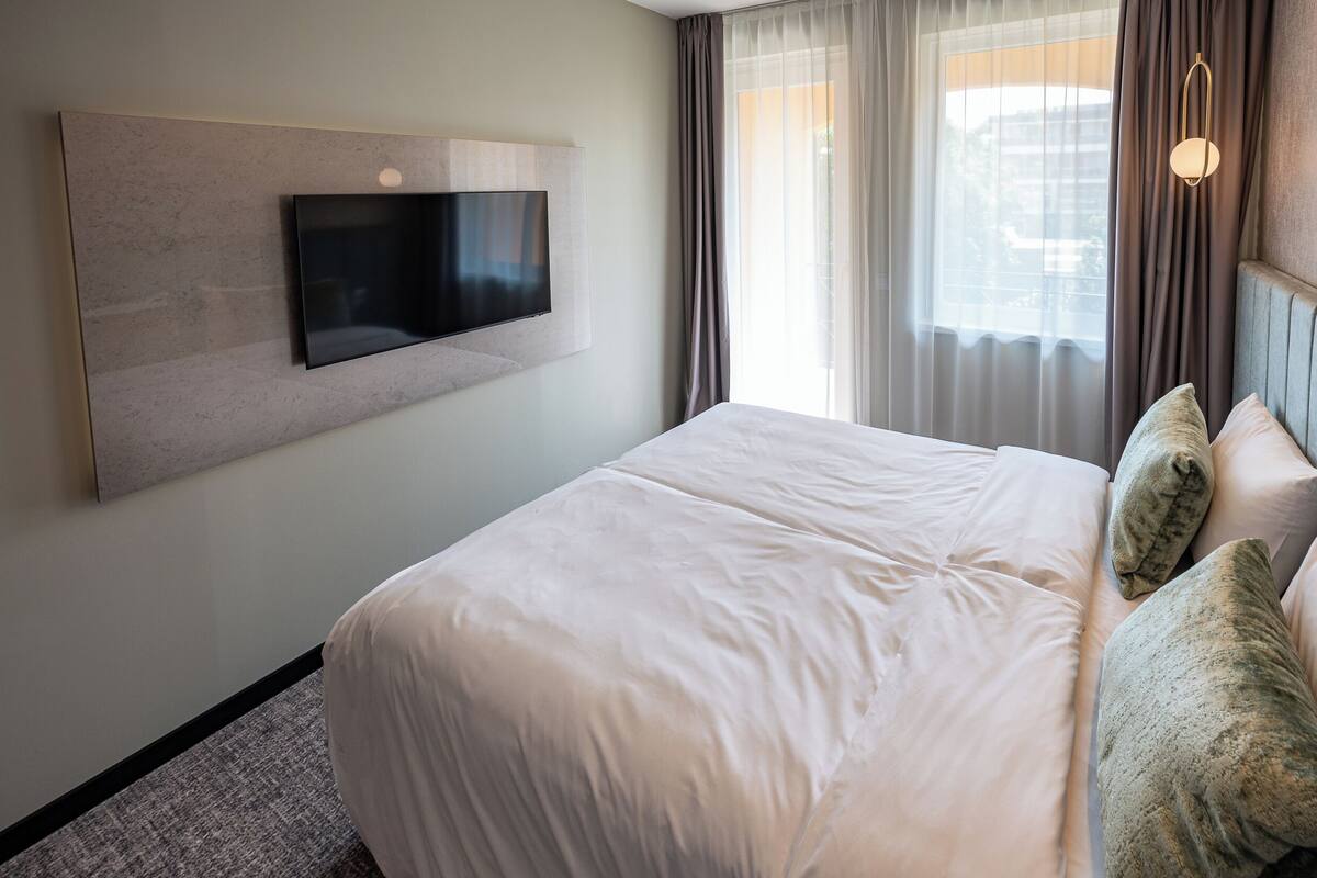 A hotel room with a large bed, a mounted TV, and a view of the city through sheer curtains.