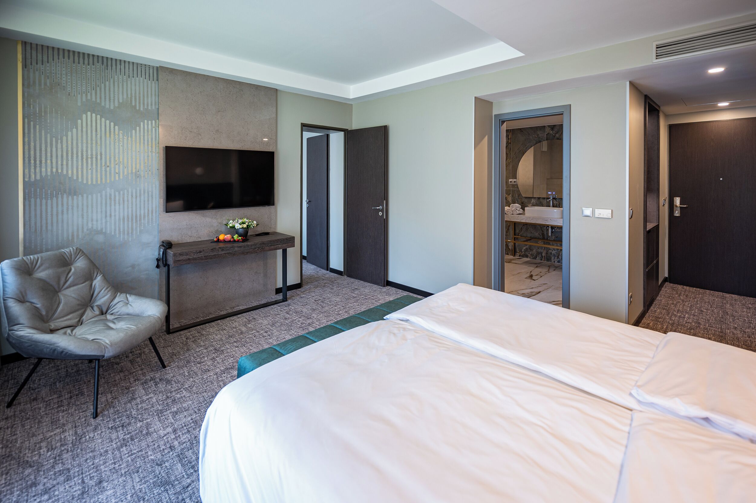 A hotel room with a large bed, a seating area with a chair, a small table with a vase, and a TV mounted on the wall.