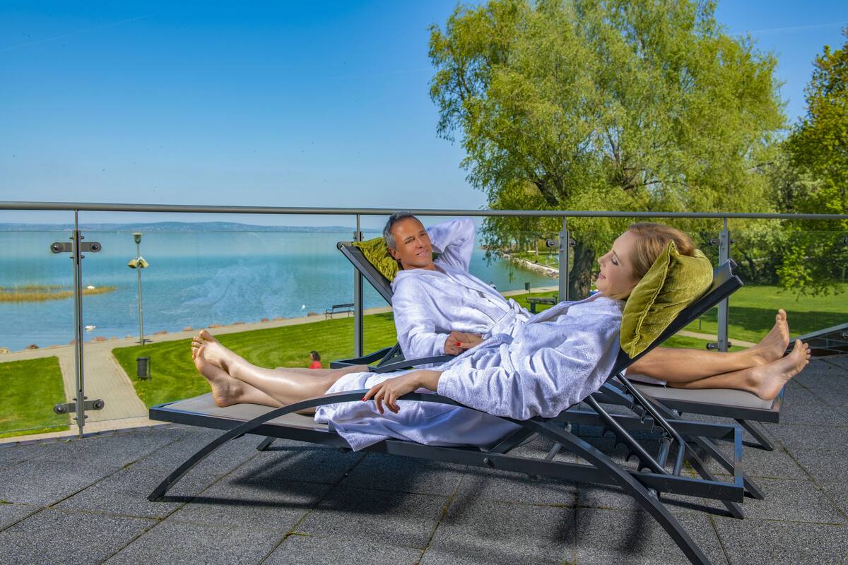 Two people relaxing on sun loungers with a lake view.