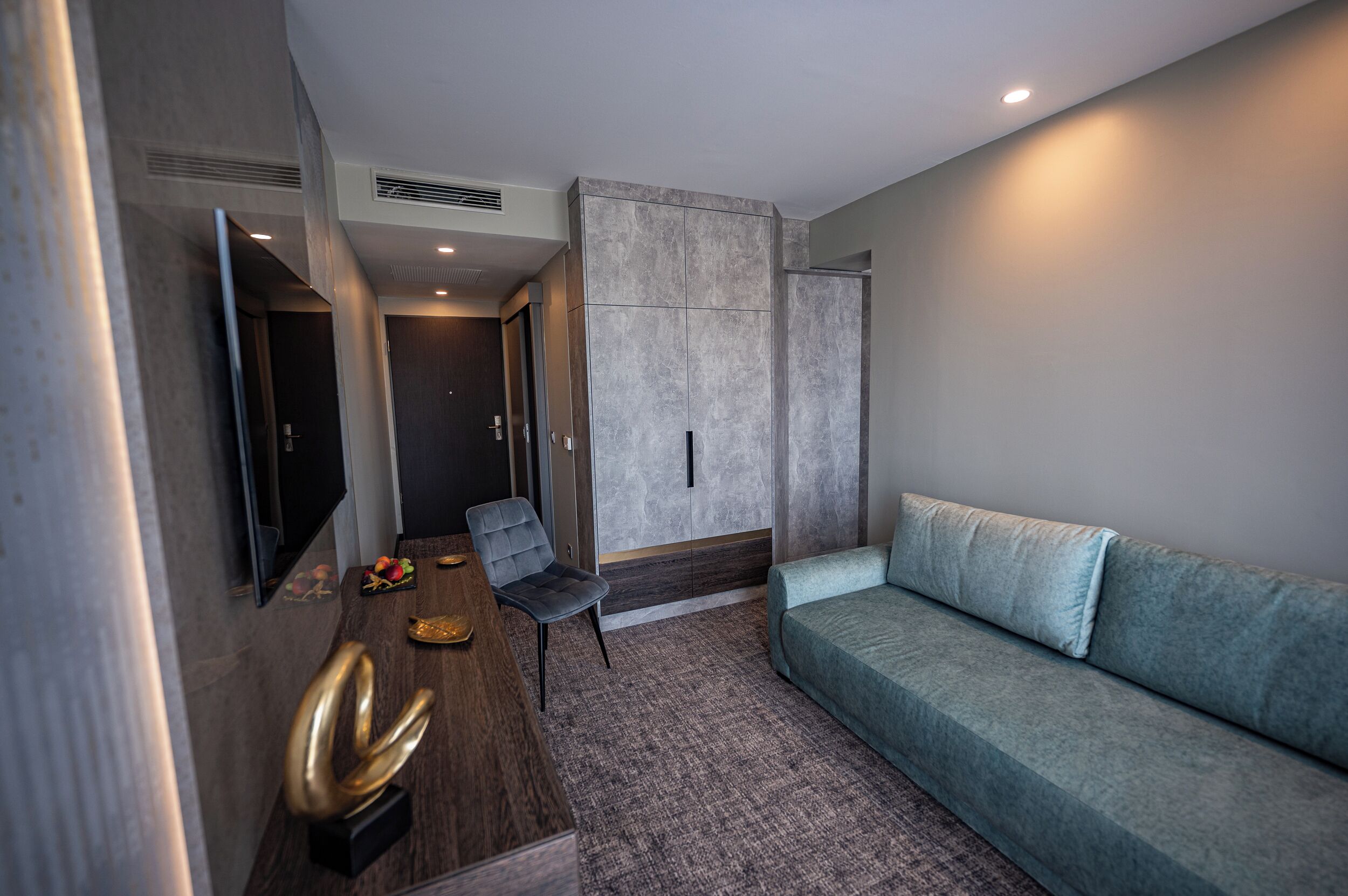 A modern hotel room with a grey sofa, a wooden side table, a flat-screen TV, and a wall-mounted cabinet.