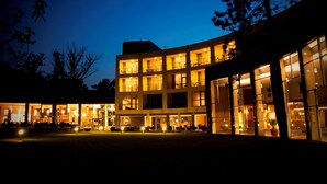 A multi-story hotel with a glass facade, outdoor seating area, and illuminated windows. - Hotel Azúr Prémium (Siófok)