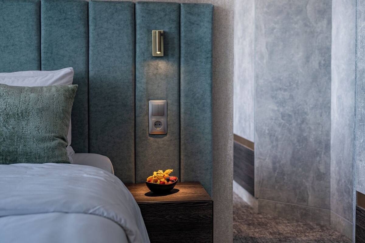 A modern hotel room with a large bed, a wall-mounted light fixture, a small table with fruit, and a textured wall.
