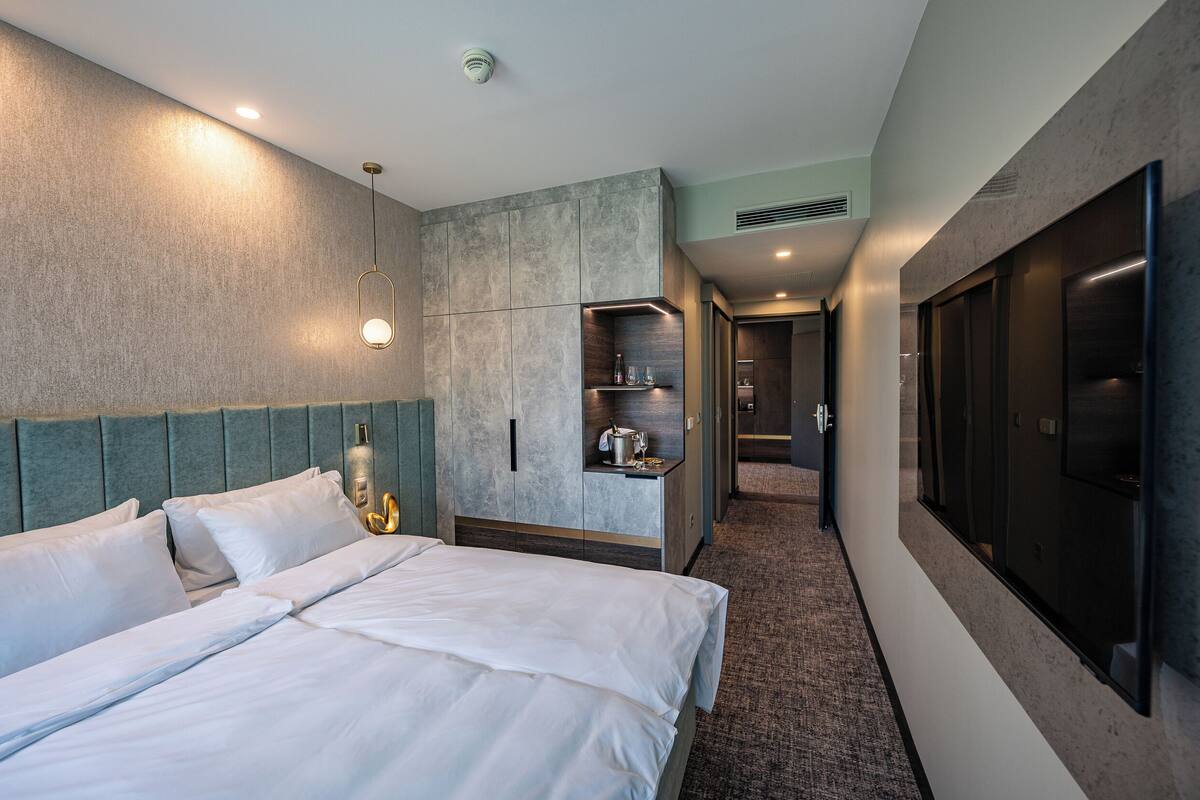 A modern hotel room with a large bed, a built-in wardrobe, a minibar, and a large TV screen.