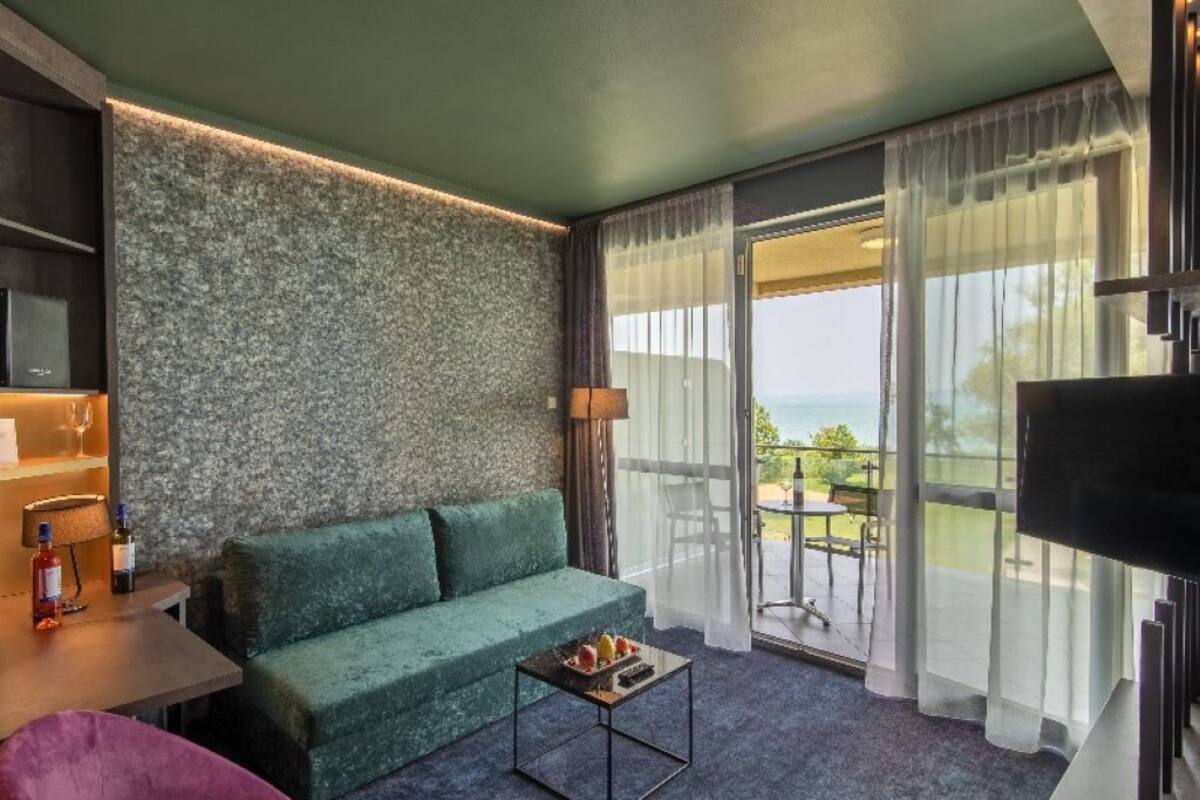 A hotel room with a green sofa, a small table with a bottle and glasses, a TV, and a view of the outdoors through large windows.