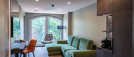 A modern living room with a green sofa, a dining area with a table and chairs, and a large window offering a view of greenery.