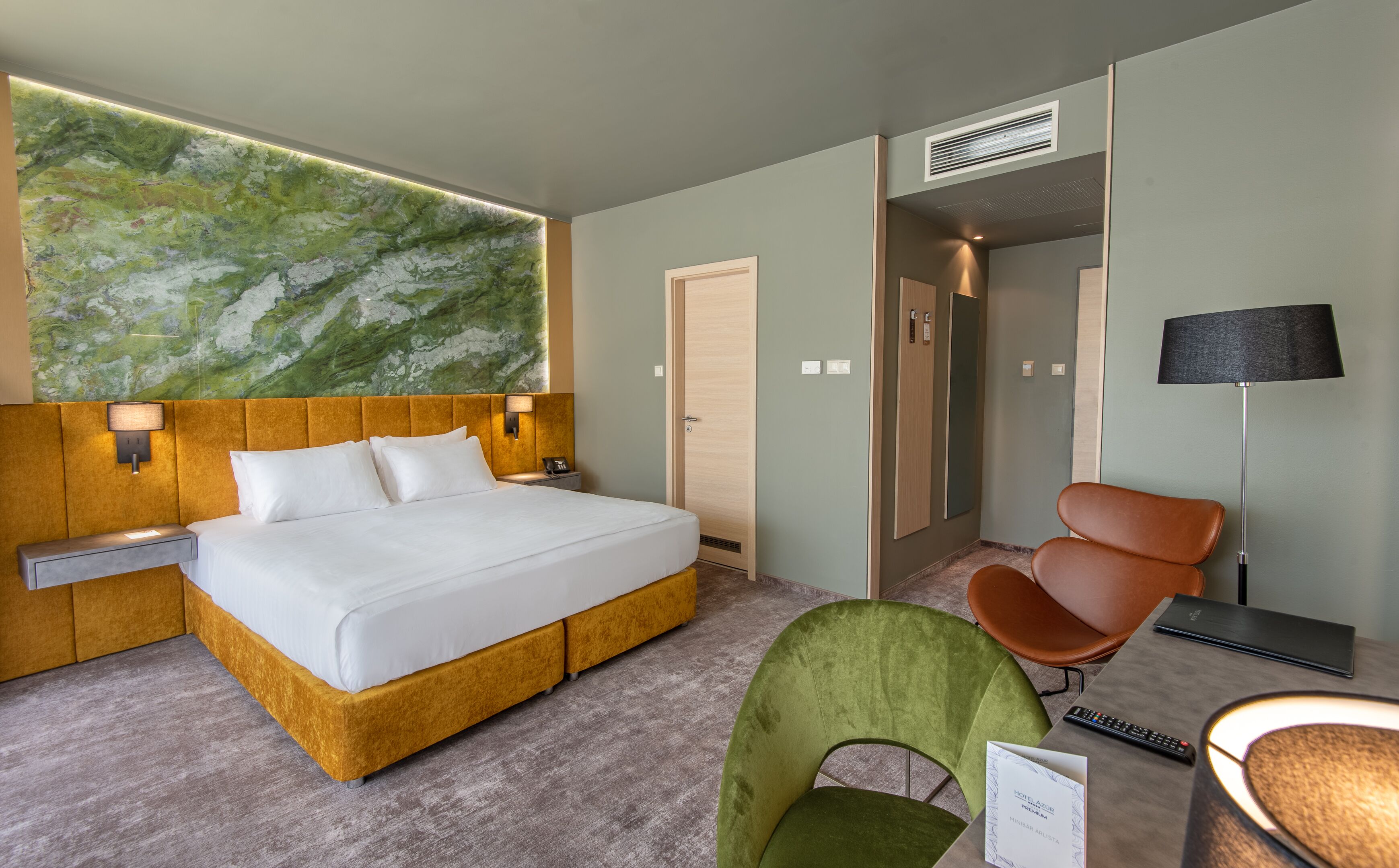 Habitación de hotel con una cama, una silla verde, una silla marrón, un escritorio, una lámpara y un cuadro abstracto verde en la pared.