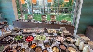 A buffet with various food items and a view of a green lawn through large windows. - Hotel Azúr Prémium (Siófok)