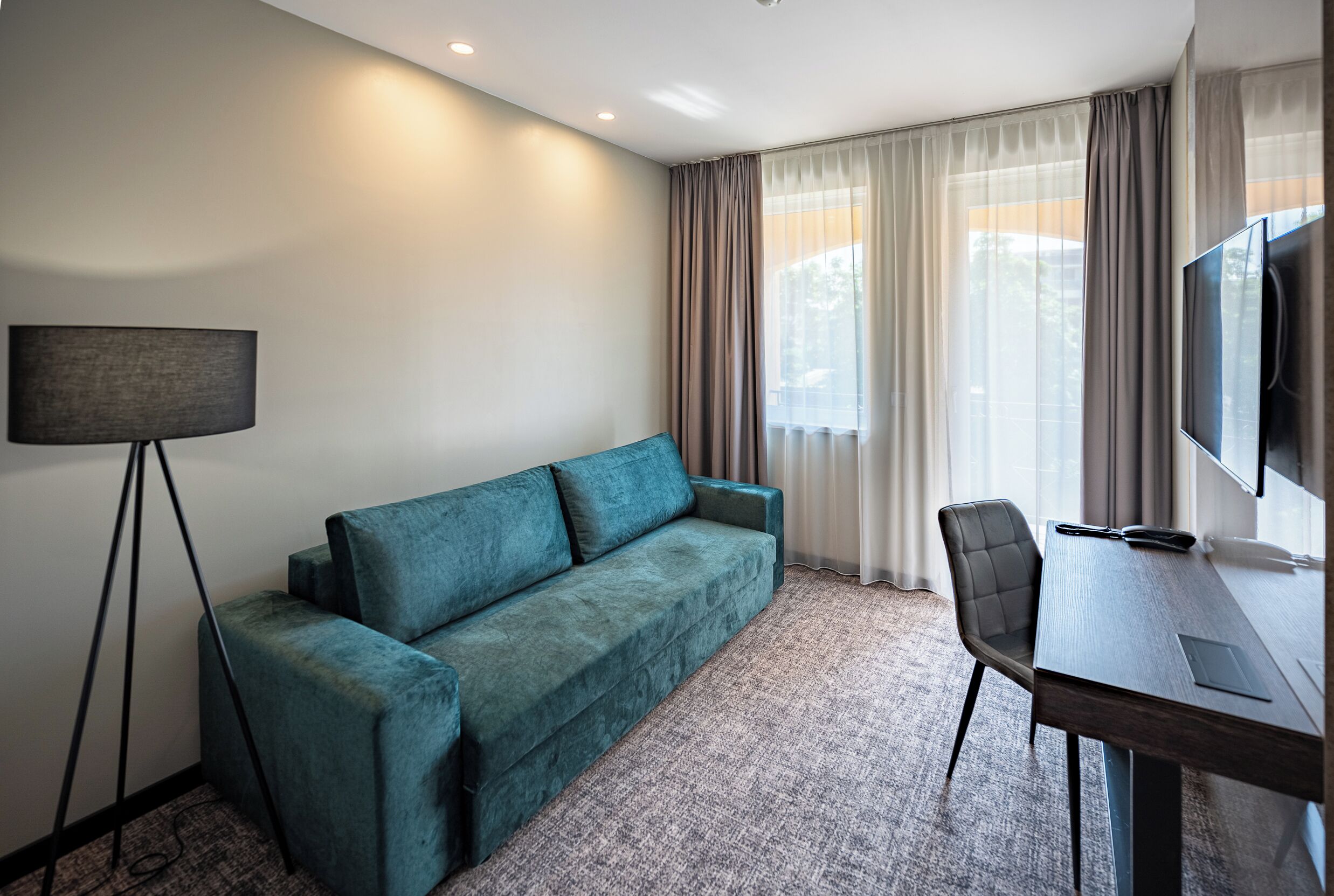 A modern hotel room with a teal sofa, a desk with a chair, a flat-screen TV, and a floor lamp.