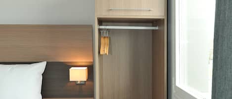 Una habitación de hotel con una cama, una mesita de noche con lámpara, un armario de madera y una ventana con cortinas.